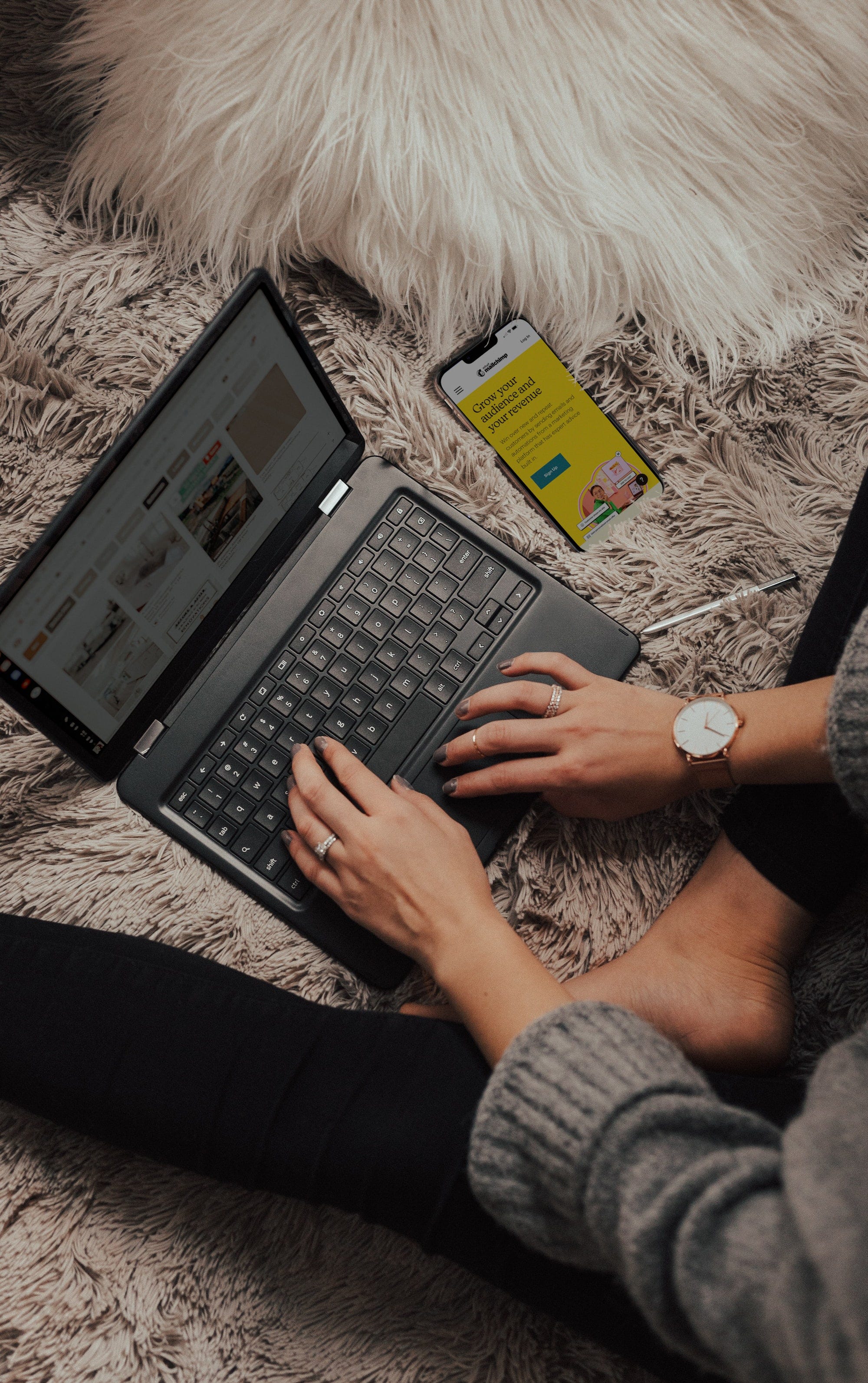 An image of a woman seated on her bed creating content on her laptop with a phone besides her