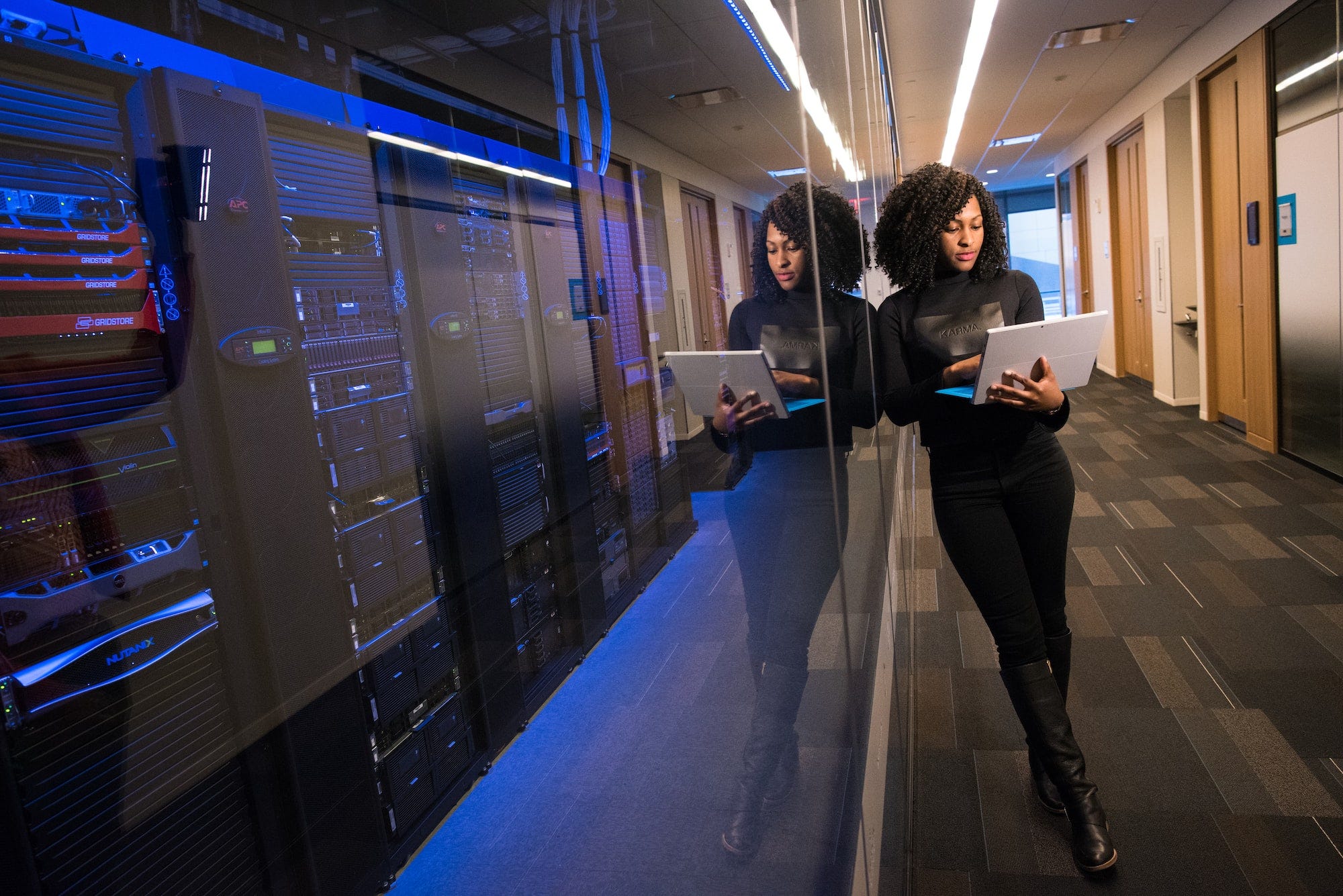 An image of a woman standing outside the glass wall of a server room with her laptop