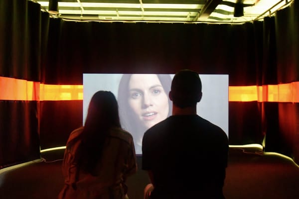 Two people, a man and a woman, mesmerized by a woman on a screen as cult members are with a cult leader